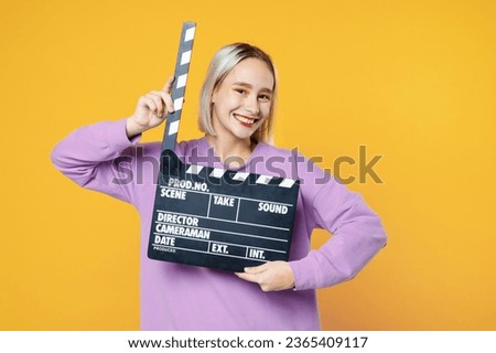 Young blonde caucasian smiling woman 20s with bob haircut bright makeup wearing casual basic purple shirt holding classic black film making clapperboard isolated on yellow background studio portrait