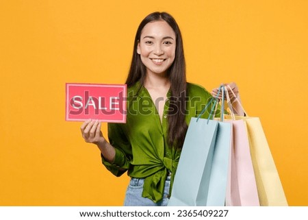 Smiling young woman of Asian ethnicity 20s wear basic green satin shirt hold package bags with purchases after shopping sign with sale title look camera isolated on yellow background studio portrait