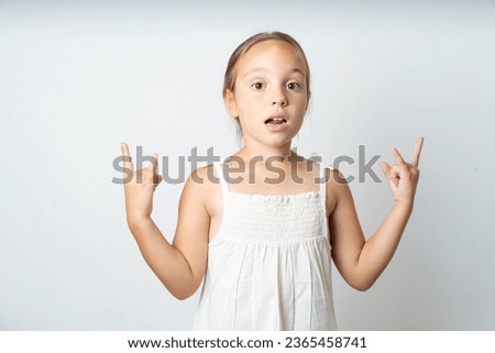 Young beautiful kid girl wearing white dress makes rock n roll sign looks self confident and cheerful enjoys cool music at party. Body language concept.