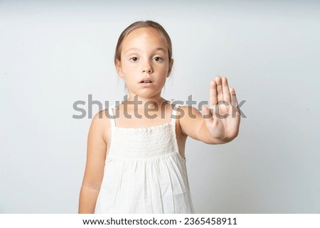 Young beautiful kid girl wearing white dress shows stop sign prohibition symbol keeps palm forward to camera with strict expression