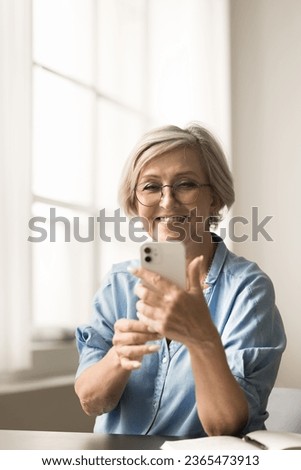 Happy retired mature lady in stylish glasses taking selfie picture on smartphone, talking on video call at work table, using Internet technology for communication, having fun