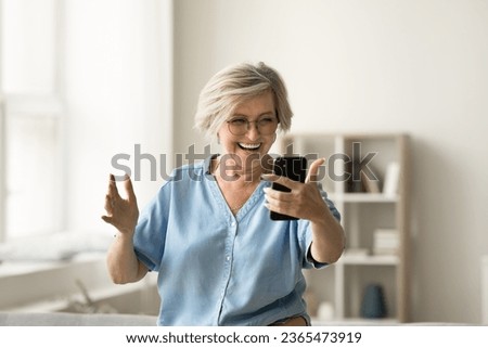 Happy excited mature older woman talking on video call on smartphone, enjoying funny conversation, laughing, taking selfie, having fun at home, using gadget for online communication Royalty-Free Stock Photo #2365473919