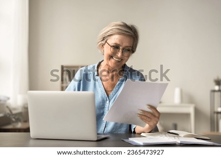 Happy mature freelance business woman in glasses reading good terms, conditions in legal document at home workplace, holding paper letter, smiling, laughing, sitting at laptop Royalty-Free Stock Photo #2365473927