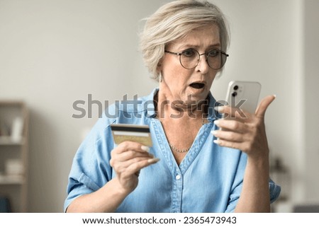 Shocked concerned mature woman in glasses holding credit card, staring at smartphone in bad surprise, getting stress, financial problems, money stealing, overspending, bankruptcy risk