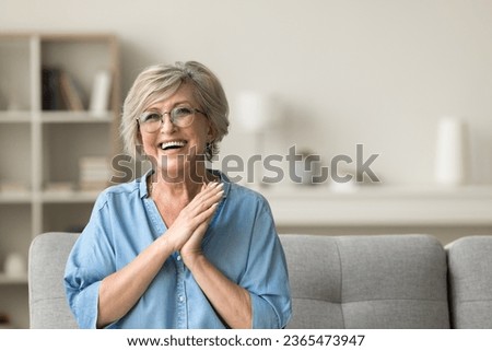 Cheerful pretty older woman in elegant glasses sitting on cozy home couch, smiling with perfect white teeth, laughing with hands at chest gesture, enjoying leisure, comfort, having fun Royalty-Free Stock Photo #2365473947