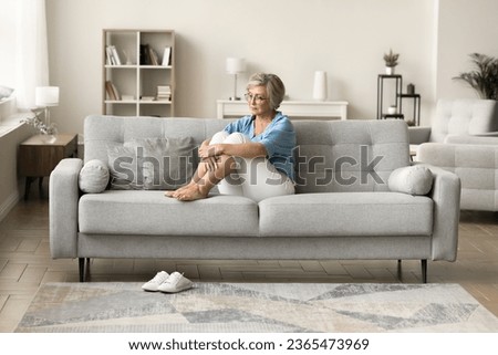 Depressed older retired woman sitting on sofa at cozy modern home, looking away in deep thoughts, staring into vacancy, thinking on depression, apathy, loneliness, remembering Royalty-Free Stock Photo #2365473969