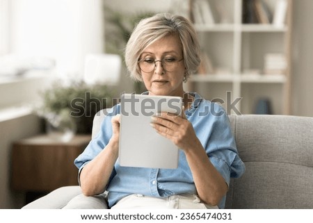 Focused blonde senior woman in glasses browsing Internet on tablet, using digital gadget, wireless technology at home, consulting doctor with medical healthcare online application