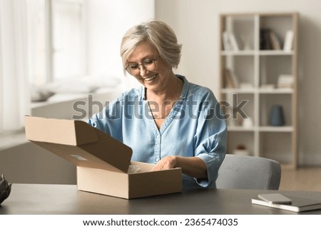 Positive satisfied customer woman receiving parcel, unpacking paper container at home, opening cardboard box with toothy smile, checking package with purchase from post transportation service Royalty-Free Stock Photo #2365474035