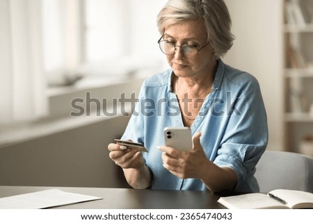 Serious senior bank client woman paying on Internet by credit card, using financial app on mobile phone for online transactions, payment, sitting at workplace table