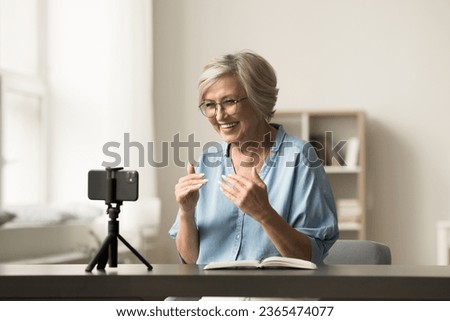 Cheerful senior elder influencer woman working on shooting for blog, recording selfie video at home, speaking at cellphone camera fixed on tripod, smiling, broadcasting online Royalty-Free Stock Photo #2365474077