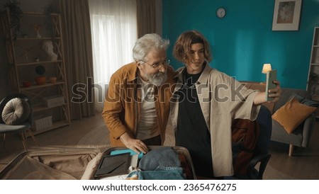 Shot of a teenager, young man having a videocall, making a video, taking a photo, selfie with his granddad helping his grandson to pack his belongings in a suitcase.