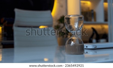 Close-up view of hourglass (sandglass or sand clock) standing on white table next to open laptop. Empty seat. Orange illumination. Soft focus. Copy space for your text. Deadline theme.