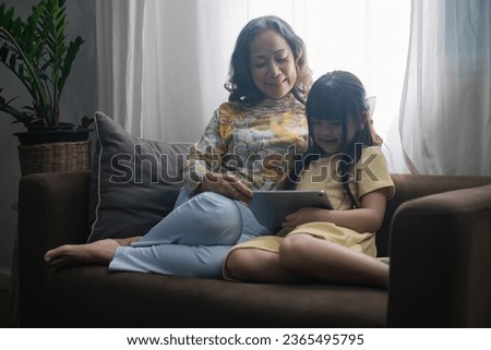 Grandma and granddaughter happily spend their free time reading books in the living room. An expression of love between an elderly woman and her adorable granddaughter.