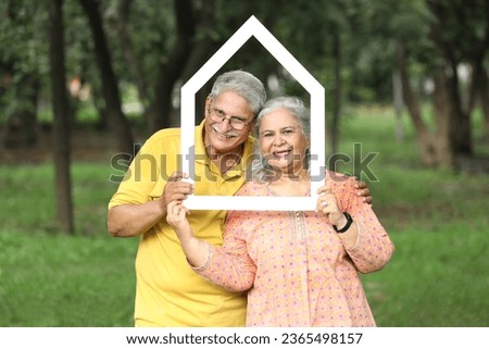 Indian happy senior couple holding Papercut Family House.. standing posing and looking in front of the camera face insert in the home shape house. Concept -  Senior lifestyle Real Estate in india. Royalty-Free Stock Photo #2365498157