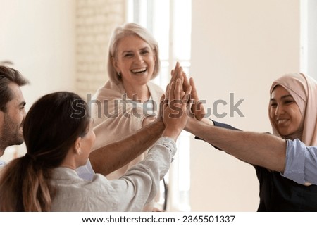 Motivated happy 50s and millennial affiliates celebrating sales growth result, common department success, taking part in team building activity, stack palm together greeting giving high five in office Royalty-Free Stock Photo #2365501337
