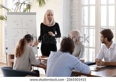 Middle eastern ethnicity coach in hijab islamic traditional wear make presentation on flip chart offer solution express opinion, explain data financial forecasting to staff, corporate training concept Royalty-Free Stock Photo #2365501415
