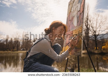 Beautiful girl with red hair and glasses painting a colorful picture on a big canvas near the lake