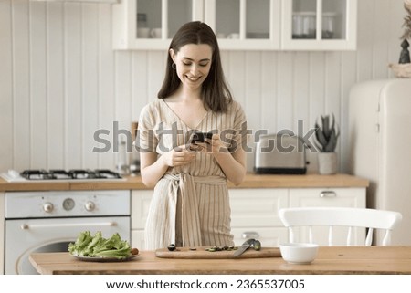 Positive adult girl browsing Internet on smartphone in kitchen, consulting cook blog, reading online recipe, typing message, preparing salad from fresh vegetables, taking picture of ingredients