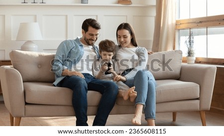 Smiling laughing young parents with cute little son using smartphone, looking at screen together, sitting on cozy couch in living room, happy family watching cartoons, shopping online at home