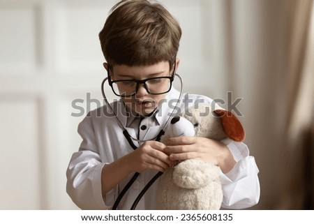 Head shot close up little boy wearing white coat uniform with stethoscope and glasses playing doctor, listening to toy, medical checkup, preschool child pretending pediatrician, children healthcare Royalty-Free Stock Photo #2365608135