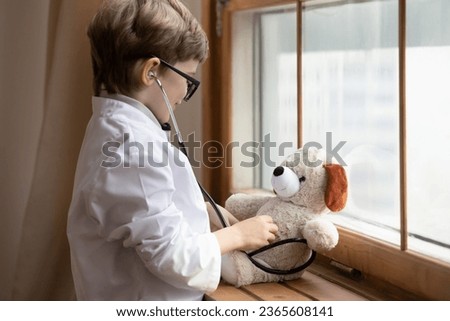 Close up cute little boy wearing white coat uniform and glasses pretending doctor, playing with fluffy toy, medical checkup, funny preschool child pretending pediatrician, children healthcare Royalty-Free Stock Photo #2365608141
