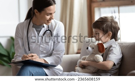 Friendly female doctor consulting little patient at meeting, writing notes in card, laughing positive preschool child hugging teddy toy, talking to smiling pediatrician, children healthcare concept