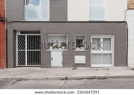 Facade of residential building with fence, white metal fence and independent loft-type house on the ground floor with pots with flowers on the windows