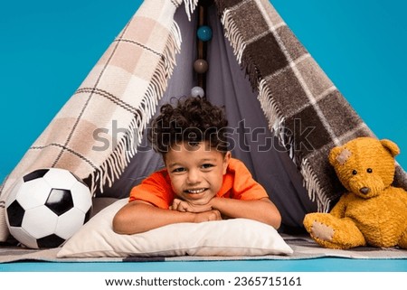 Photo of cute boy kid having nap in diy tent playing with toys lying isolated blue color background