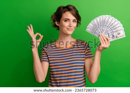 Photo portrait of attractive young woman show okey symbol hold money fan wear trendy striped clothes isolated on green color background