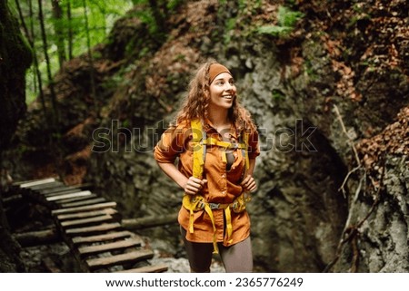 Happy woman in hiking clothes with a yellow backpack walks along a wooden hiking path in the mountains. Hiking, active lifestyle. Outdoor adventure concept. Royalty-Free Stock Photo #2365776249