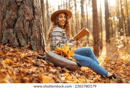 A tourist female in a hat sits in the autumn forest on yellow leaves and reads a book. Beautiful woman enjoying sunny autumn weather. Royalty-Free Stock Photo #2365780179