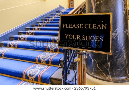 Sign saying "Please clean your shoes" at the bottom of a flight of stairs in a large, ornate building.