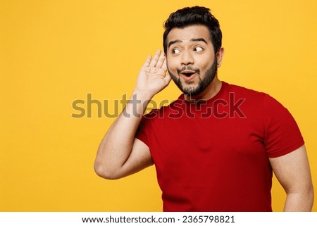 Young curious nosy happy Indian man he wearing red t-shirt casual clothes trying to hear you overhear listening intently isolated on plain yellow orange background studio portrait. Lifestyle concept