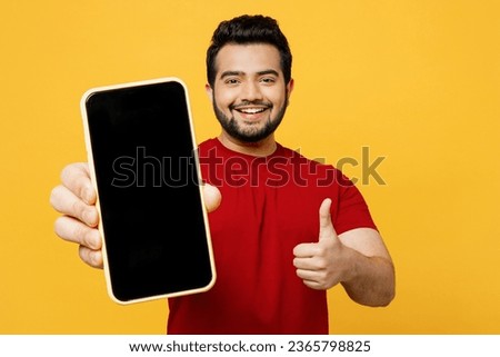 Satisfied happy young Indian man wearing red t-shirt casual clothes hold in hand use mobile cell phone with blank screen workspace area show thumb up isolated on plain yellow orange background studio Royalty-Free Stock Photo #2365798825