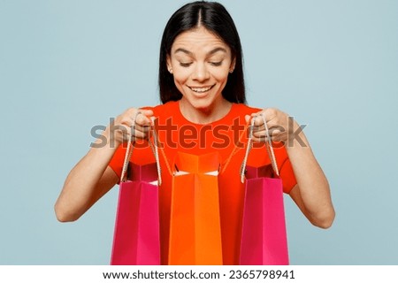 Young latin woman wearing orange red t-shirt casual clothes hold in hand open paper package bags after shopping isolated on plain light pastel blue cyan background. Black Friday sale buy day concept