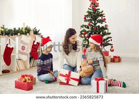 Family opening christmas presents. Happy mother with young son and daughter children opens gift box on Christmas morning. Mom and kids in santa hats sitting on floor near Christmas tree at home.