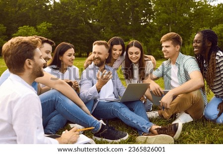 Group of happy, cheerful, diverse, multiethnic student friends sitting on green grass outside, studying information for project together, discussing something, using online tools on modern laptop PC Royalty-Free Stock Photo #2365826001