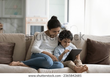 Smiling Indian mother with son using tablet at home, relaxing sitting on cozy couch together, loving young mom and 5s boy child looking at gadget screen, watching video or cartoons, playing games
