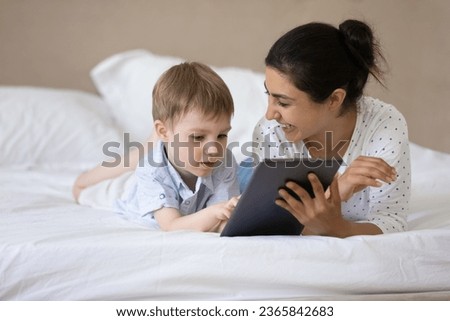 Happy Indian mother with 7s son using tablet together, relaxing lying on cozy bed at home, smiling caring young mom and adorable boy child having fun with device, watching cartoons, enjoying weekend