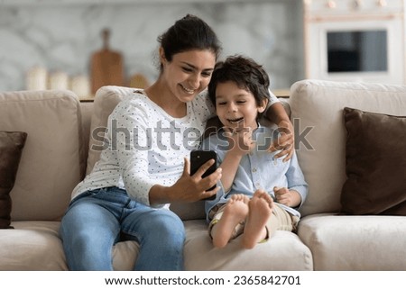 Smiling Indian mother and son having fun with smartphone together, hugging, sitting on cozy sofa, happy young mom with 5s boy child looking at phone screen, watching cartoons or making video call