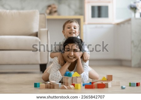 Portrait happy Indian mother with Caucasian son having fun, playing with toys on warm floor at home, smiling 7s boy sitting on young mom back, looking at camera, family enjoying leisure time Royalty-Free Stock Photo #2365842707