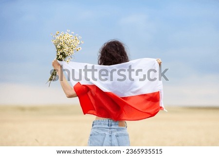 Woman with bouquet of daisies holding flag of Poland in wheat field. Back view. Polish Flag Day. Independence Day. Travel and learn polish language concept. Royalty-Free Stock Photo #2365935515