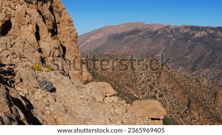 Walking on the mountain slope with large orange or red rocks on the path, little vegetation, dry, torrid, in the Atlas of Morocco, with bushes, intense physical effort, in full sun