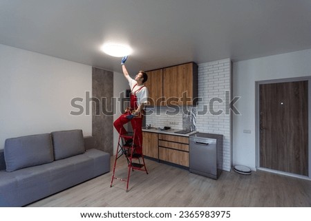 Worker installing lamp on stretch ceiling indoors. Royalty-Free Stock Photo #2365983975