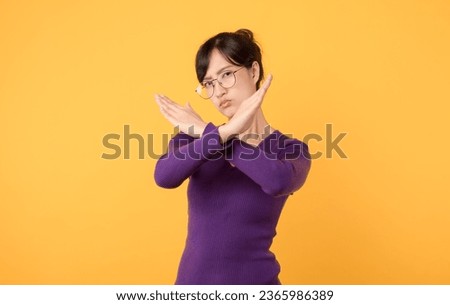 Explore concept of denial and rejection through X gesture made serious young woman wearing purple shirt and eyeglasses in studio portrait against a yellow background. impactful symbol for caution.