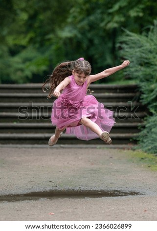 A little girl in a beautiful pink dress with long hair runs quickly and jumps over a puddle. I concentrated and bit my tongue. Outdoor games. Blurred background.