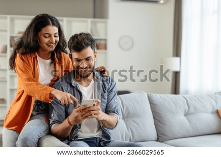 Happy young indian couple using smartphone, wife pointing at cellphone screen, sitting on sofa in living room interior at home. New app ad, banner Royalty-Free Stock Photo #2366029565