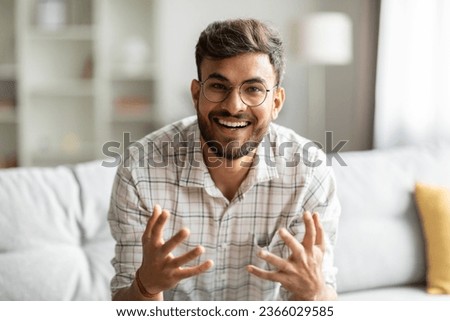 Cheerful indian guy talking looking at camera and gesturing, communicating online sitting on couch at home. Web camera view of man chatting remotely via video call Royalty-Free Stock Photo #2366029585