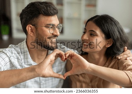 Portrait of young romantic indian couple making heart gesture, looking at each other and smiling, spouses connecting their hands together into love sign