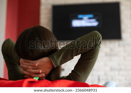 Back view photo of brunette young woman in casual clothes sitting on couch in living room, holding remote, switching channels on TV, weekend at home, rest and relax